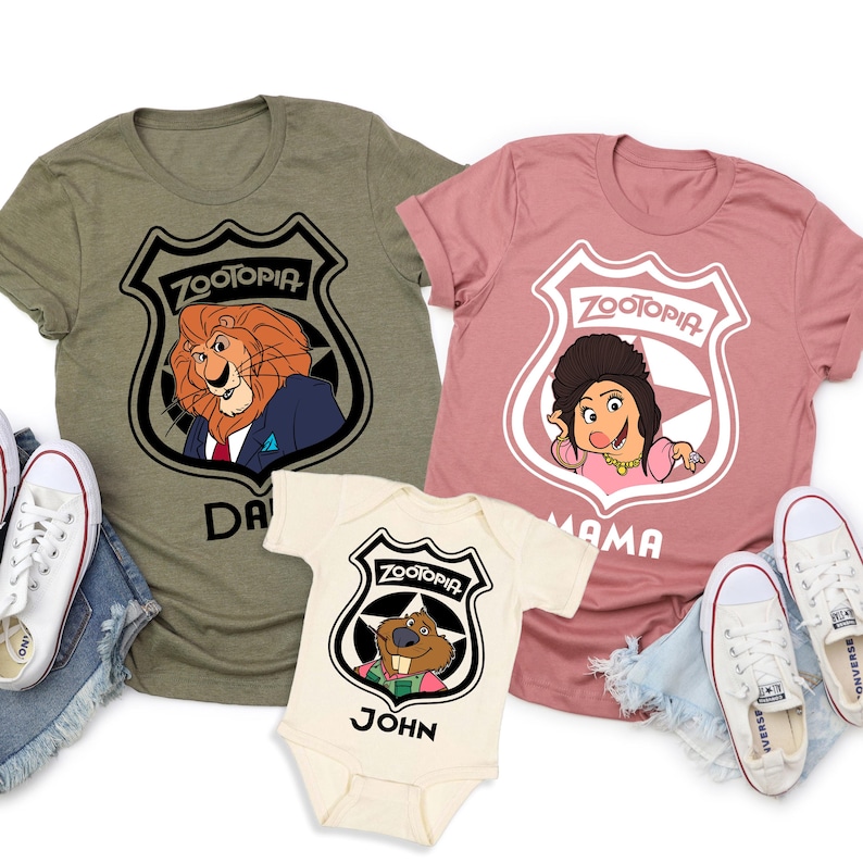 May include: Three Zootopia-themed t-shirts and a onesie. The shirts are olive green and dusty rose, and the onesie is cream-colored. Each features a Zootopia badge with a character image and a name. White sneakers are visible on either side of the shirts.