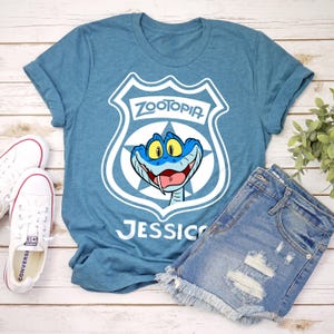 May include: A teal blue t-shirt featuring a white graphic of a Zootopia police badge with a blue snake character. The name "Jessica" is printed below the graphic. The shirt is paired with denim shorts and white Converse sneakers.