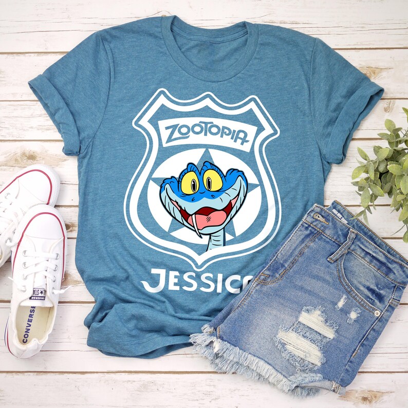 May include: A teal blue t-shirt featuring a white graphic of a Zootopia police badge with a blue snake character. The name "Jessica" is printed below the graphic. The shirt is paired with denim shorts and white Converse sneakers.