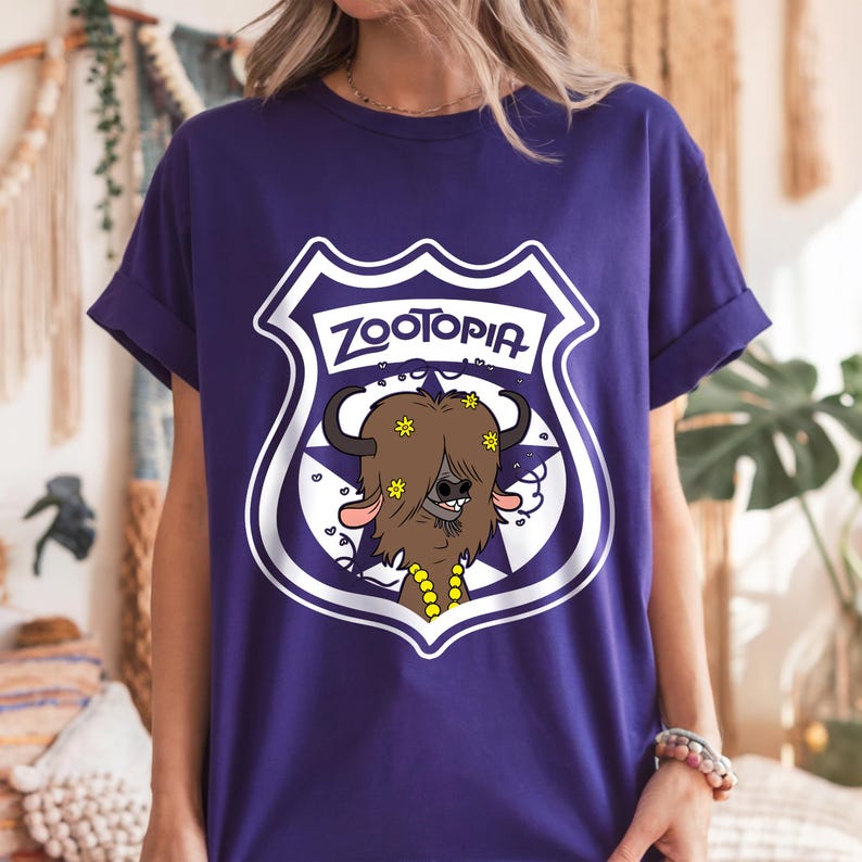 May include: A purple t-shirt featuring a Zootopia police badge design. The badge includes the word "Zootopia" and a cartoon bison character with yellow flower accents. The shirt has rolled-up sleeves.
