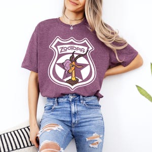 May include: A maroon t-shirt featuring a white shield design with the word "ZOOTOPIA" and a cartoon gazelle character. The gazelle has purple hair and is wearing a brown top. The shirt is paired with blue jeans.