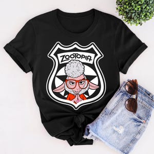 May include: Black t-shirt featuring a graphic from the movie Zootopia. The design includes a sheep character wearing glasses, set within a police badge shape, with the word "Zootopia" at the top. The shirt is made of a soft material.