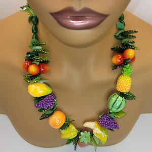 Könnte beinhalten: Eine farbenfrohe Halskette mit verschiedenen Fruchtanhängern, darunter Tomaten, Mangos, Trauben, Orangen und eine Ananas. Die Halskette besteht aus grünen Blättern und Perlen, mit einer Banane und einer Erdbeere. Die Halskette wird auf einer Schaufensterpuppe präsentiert.