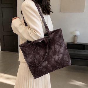 May include: A dark charcoal gray tote bag featuring a textured, distressed finish with a geometric stitched pattern. The bag has two shoulder straps and a rectangular shape, constructed from a matte material with visible seam detailing throughout.