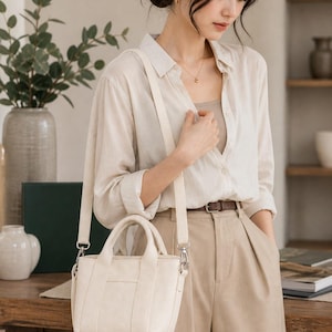 Può includere: Borsa a mano color crema con manico superiore e tracolla staccabile. La borsa ha una forma rettangolare con dettagli di cucitura verticali. La donna indossa una camicia beige abbottonata e pantaloni color cachi.