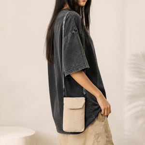 Könnte beinhalten: Eine beige Umhängetasche für das Telefon mit Klappenverschluss hängt an einem schwarzen Riemen. Die Tasche wird über einem schwarzen T-Shirt und khakifarbenen Shorts getragen. Eine braune Baseballkappe vervollständigt das lässige Outfit.