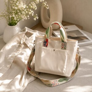 May include: Cream-colored tote bag with a floral patterned strap and handles. The bag has a front pocket and silver hardware. A white shirt and vase of flowers are in the background.