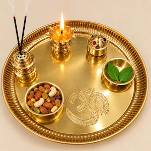 May include: A round brass puja thali plate featuring an embossed Om symbol. The tray holds a lit oil lamp, an incense stick holder with two burning incense sticks, a small lidded container, a bowl of mixed nuts, and a bowl with green leaves.