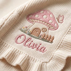 May include: A cream-coloured knit jumper featuring a ruffled edge and an embroidered design of a mushroom house with a pink cap, a small butterfly, and flowers. The name Olivia is embroidered in pink cursive script below the mushroom house.