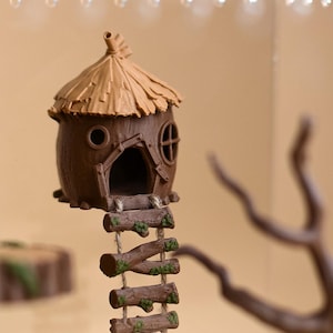 Pode incluir: Uma cabana miniatura castanha com telhado de palha texturizado, uma janela circular e uma entrada em arco. Uma escada de corda com degraus de madeira horizontais com detalhes de musgo verde pende da base e desce verticalmente.