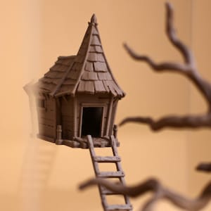 Pode incluir: Uma pequena casa na árvore de madeira marrom com um telhado pontudo e uma escada que leva à entrada. A casa na árvore está situada em um fundo desfocado com um galho de árvore em primeiro plano.