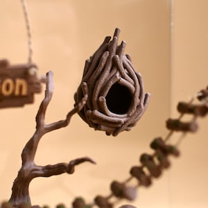 May include: A brown decorative birdhouse, resembling a nest, hangs from a branch. The birdhouse features a circular entrance and is constructed from interwoven twigs. A sign with the word "sion" is visible.