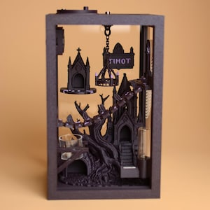 May include: A rectangular black display box containing a 3D-printed gothic scene with a dark tree, a small chapel, a hanging chandelier, and a sign with the text TIMOT. The interior features miniature architectural elements and a clear glass cup.