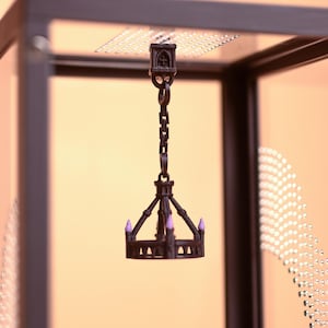 Può includere: Un lampadario in miniatura color marrone scuro con una struttura circolare e bracci multipli, ognuno sormontato da un piccolo dettaglio a forma di candela color viola chiaro. Il lampadario è sospeso tramite una catena a un supporto quadrato.