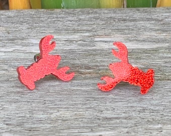 May include: A pair of red glitter lobster earrings. The earrings are shaped like lobsters and have a shiny, glittery finish.