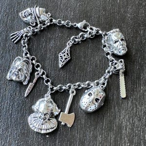 May include:  Stainless steel silver charm bracelet with various horror movie character charms. The charms include a clown, a doll, a hockey mask, a chainsaw, an axe, and a knife. The bracelet is on a dark graysurface.