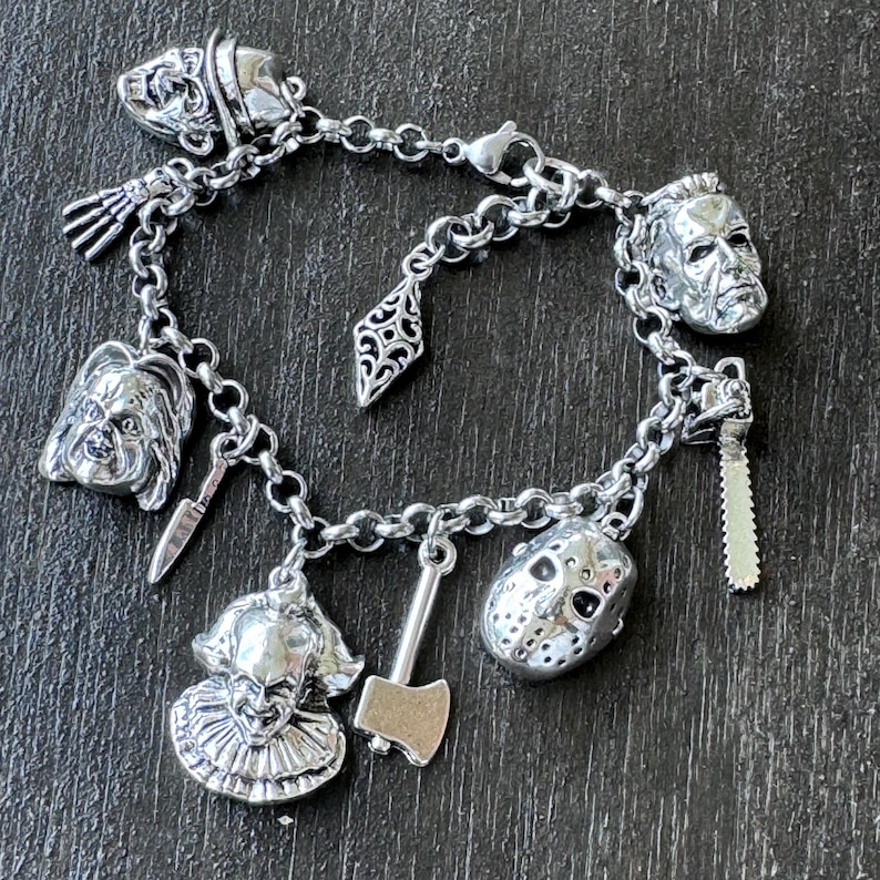 May include:  Stainless steel silver charm bracelet with various horror movie character charms. The charms include a clown, a doll, a hockey mask, a chainsaw, an axe, and a knife. The bracelet is on a dark graysurface.