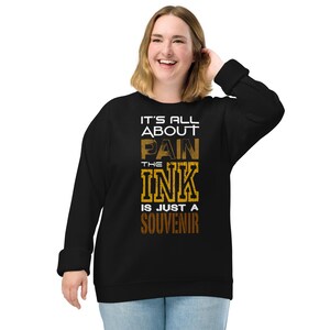 Könnte beinhalten: Schwarzes Sweatshirt mit dem Text "IT'S ALL ABOUT PAIN THE INK IS JUST A SOUVENIR" in weißer, brauner und goldener Schrift. Das Sweatshirt hat lange Ärmel und einen Rundhalsausschnitt.