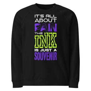 Könnte beinhalten: Schwarzes Sweatshirt mit dem Text "IT'S ALL ABOUT PAIN THE INK IS JUST A SOUVENIR" in Weiß, Lila und Grün. Das Sweatshirt hat einen Rundhalsausschnitt und lange Ärmel. Der Text ist fett und im Used-Look.
