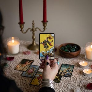 May include: A hand holds a tarot card with a yellow background and a figure in colorful clothing. Other tarot cards are laid out on a lace-covered surface, along with candles, a brass candelabra, and a wooden bowl of green stones. The card reads "THE FOOL."