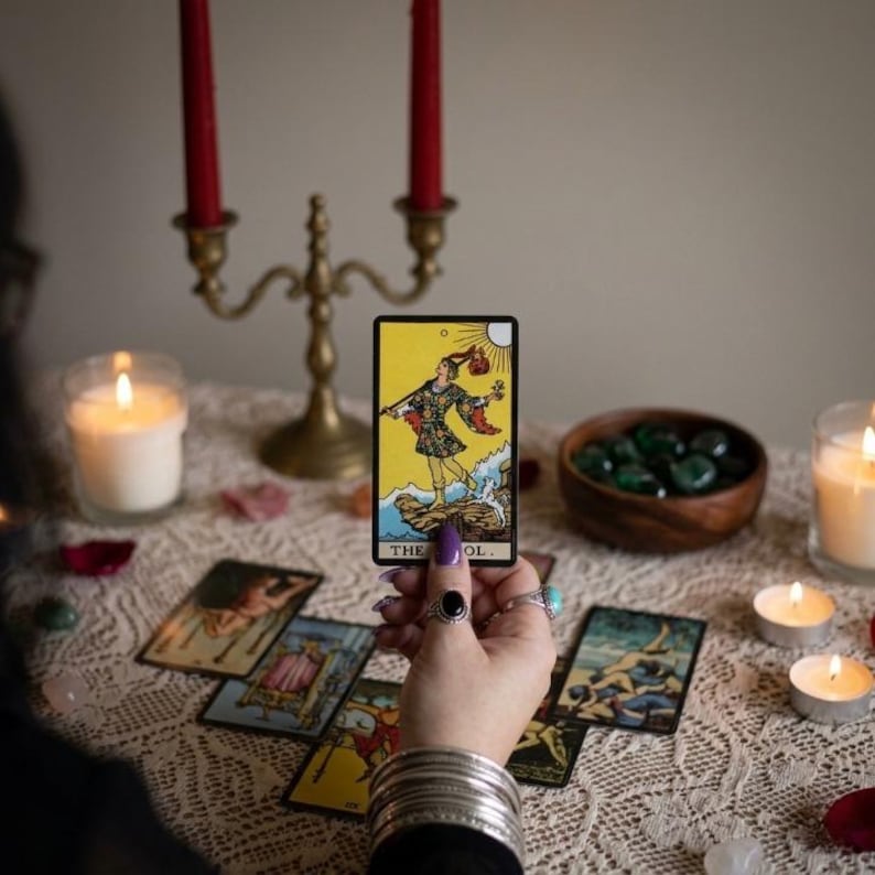 May include: A hand holds a tarot card with a yellow background and a figure in colorful clothing. Other tarot cards are laid out on a lace-covered surface, along with candles, a brass candelabra, and a wooden bowl of green stones. The card reads "THE FOOL."