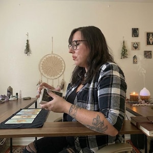 May include: A person wearing glasses and a black and white plaid shirt is holding a deck of tarot cards. Several cards are laid out on a black mat on a wooden table. A dream catcher and other decorative items are on the wall.