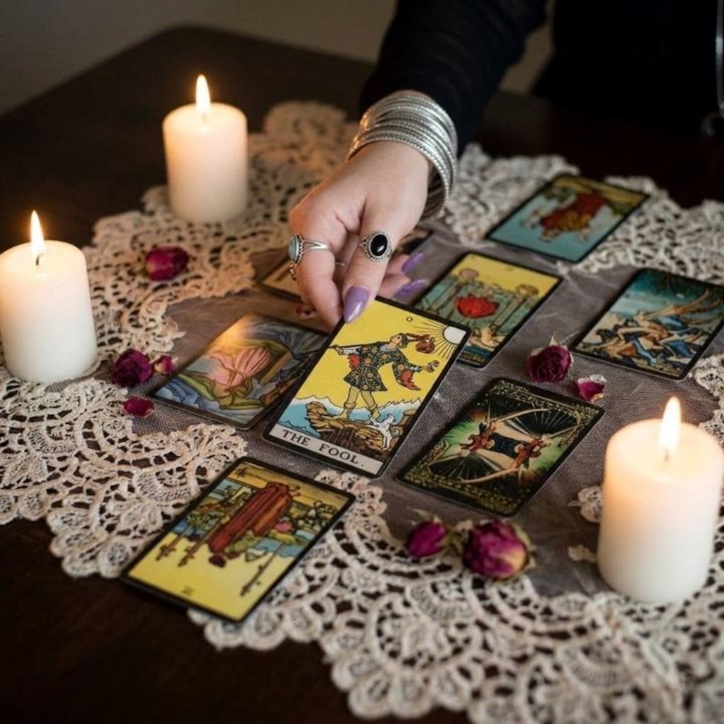 May include: A tarot card reading setup with lit candles, dried rosebuds, and a hand holding a card labeled "THE FOOL." The cards feature colorful illustrations. The scene is set on a white lace tablecloth, creating a mystical atmosphere.