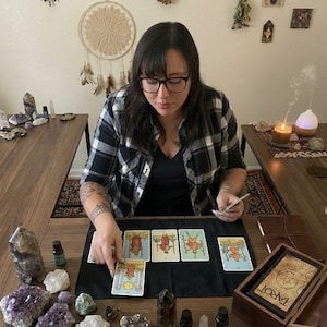 May include: A person wearing glasses and a plaid shirt is seated at a wooden table, arranging tarot cards. The table is adorned with crystals, essential oil bottles, and a wooden box labeled "TAROT". A lit candle and a diffuser are visible.