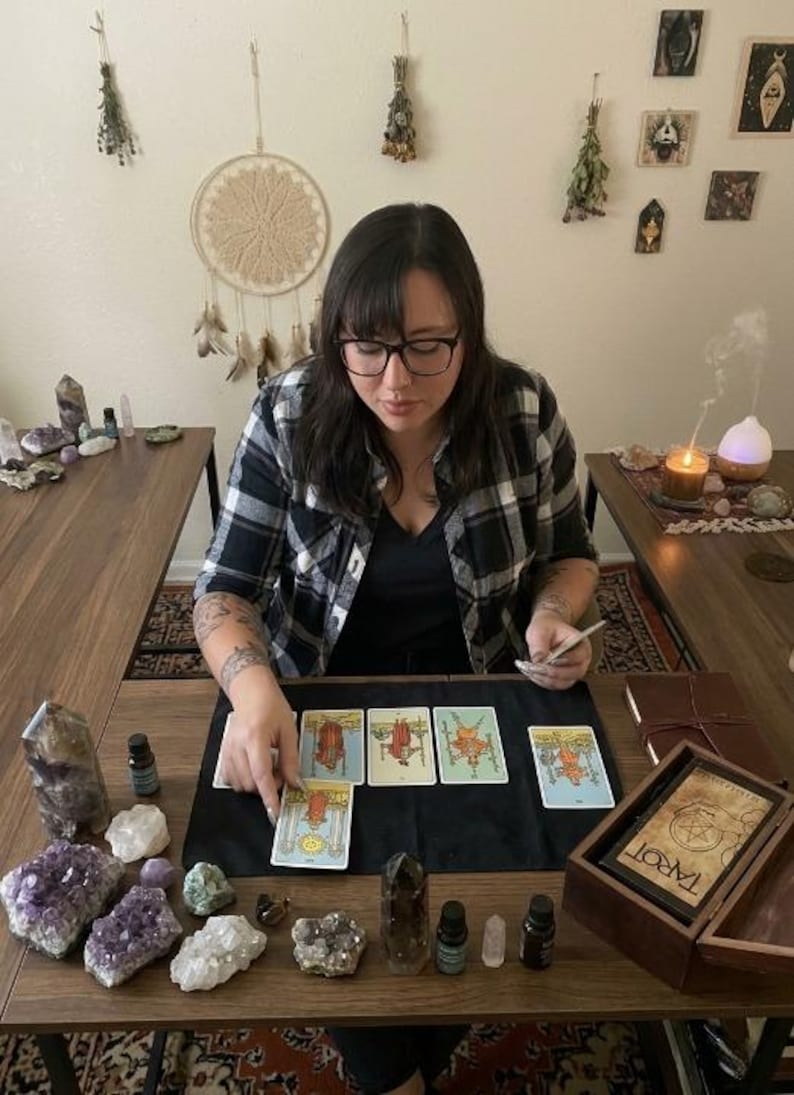 May include: A person wearing glasses and a plaid shirt is seated at a wooden table, arranging tarot cards. The table is adorned with crystals, essential oil bottles, and a wooden box labeled "TAROT". A lit candle and a diffuser are visible.