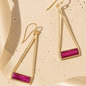 May include: A pair of gold-colored earrings with a triangular design. Each earring features a rectangular, magenta-colored gemstone set within the lower portion of the triangle. The earrings are displayed on a cream-colored surface with a satin ribbon.