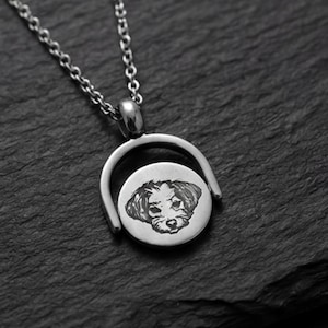 Puede incluir: Un collar con colgante de metal plateado que presenta un dije circular suspendido dentro de un marco semicircular. El dije circular muestra un grabado oscuro y detallado de la cara de un perro en su superficie frontal, unido a una cadena fina.