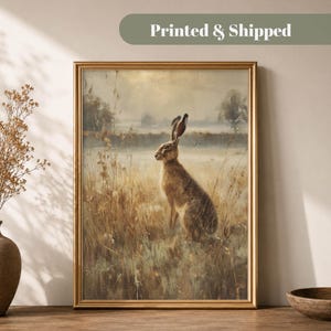 May include: A framed wall art print featuring a brown hare standing in a field of tall, dry grasses. The scene is set against a muted, hazy landscape with soft, neutral tones, captured in a painterly style within a gold-colored frame.