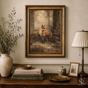 May include: A framed wall art print featuring a red fox standing in a misty forest, displayed above a wooden surface with a ceramic vase, a stack of three books titled The Kinfolk Home, Made for Living, and Live Beautifully, and a lamp.