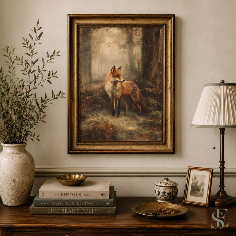 May include: A framed wall art print featuring a red fox standing in a misty forest, displayed above a wooden surface with a ceramic vase, a stack of three books titled The Kinfolk Home, Made for Living, and Live Beautifully, and a lamp.