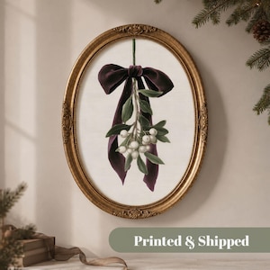 May include: An oval-shaped gold frame containing a print of a sprig of white mistletoe berries and green leaves tied with a large, dark purple velvet ribbon. The artwork is centred on a light, textured off-white background within the frame.