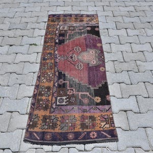 May include: A rectangular runner rug featuring a central medallion in shades of deep purple, magenta, and black. The border is decorated with repeating floral motifs in orange, brown, and dark blue, with a fringed edge at the top and bottom.
