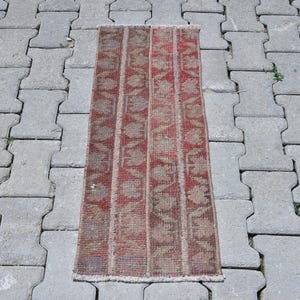May include: A rectangular woven rug featuring a repeating vertical striped pattern in shades of faded red, muted brown, and grey. The textile displays a distressed, weathered texture with visible fringe detailing along the top and bottom edges.