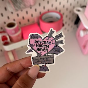 Könnte beinhalten: Ein gestanzter Aufkleber mit einem rosa Herz, das von drei grauen Pfeilen durchbohrt wird. Der Text lautet "Open REVERSE HAREM HAVEN" und "Where every room has a King...or three" mit einem kleinen Schild, das auf "ROOM FOR ALL" zeigt.