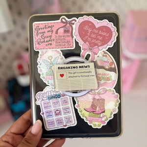 Peut inclure: Une coque de tablette noire recouverte de divers autocollants roses et blancs. Les autocollants présentent du texte incluant Greetings from my Spicy Romance era, Buy me books & tell me I'm pretty, et un graphique de journal avec le texte Breaking News This girl is emotionally attached to fictional men.