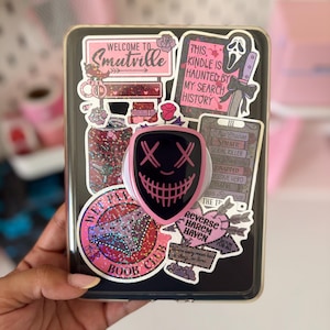 Könnte beinhalten: Eine schwarze E-Reader-Hülle, die mit verschiedenen rosa und schwarzen Aufklebern bedeckt ist, darunter Texte wie Welcome to Smutville, This Kindle is haunted by my search history und Reverse Harem Haven. Ein rosa-schwarzer, schildförmiger Handygriff ist mittig platziert.