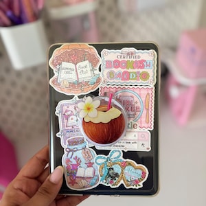 Puede incluir: Una funda transparente para tableta decorada con varias pegatinas, incluyendo una concha con un libro abierto, una bebida de coco con una flor, un frasco con vida marina y texto que dice Certified Bookish Baddie, Summer Girl Book Club y Enemies to Lovers.