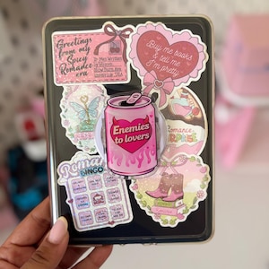 Peut inclure: Une coque de tablette rectangulaire noire recouverte d'autocollants roses, incluant un support central en forme de canette de soda avec l'inscription Enemies to lovers, une carte de bingo Romance et un autocollant en forme de cœur Buy me books.