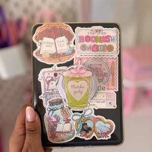 Peut inclure: Un étui de tablette rectangulaire noir décoré de divers autocollants, dont un graphique de club de lecture en forme de coquillage, une étiquette Certified Bookish Baddie, un bocal de trésors marins et un support central transparent.