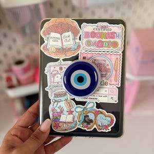 Peut inclure: Une coque de tablette transparente décorée de divers autocollants, dont un coquillage avec un livre ouvert, un support œil bleu, un bocal rempli de vie marine et des textes comme Certified Bookish Baddie et Girl Dinner avec une barre de recherche.