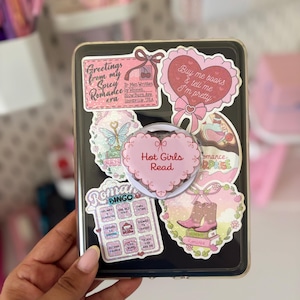 Op de afbeelding: Een zwarte tablethoes versierd met diverse stickers, waaronder een hartvormige telefoonhouder met de tekst Hot Girls Read. Andere stickers bevatten teksten zoals Greetings from my Spicy Romance era en Romance Bingo op de achterkant.