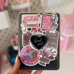 Peut inclure: Une liseuse noire recouverte d'autocollants roses et noirs, avec des textes comme Welcome to Smutville, This Kindle is haunted by my search history et Wet Panty Book Club. Un support de téléphone noir en forme de cœur avec Good girl.