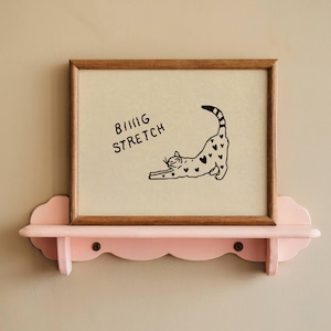 Puede incluir: Una impresi&oacute;n enmarcada con un marco de madera marr&oacute;n que presenta un gato negro estir&aacute;ndose con marcas en forma de coraz&oacute;n. La impresi&oacute;n tiene las palabras "BIG STRETCH" en negro. La impresi&oacute;n se muestra en una estanter&iacute;a rosa en forma de nube.