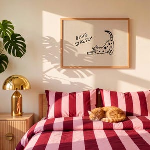 Puede incluir: Un estampado enmarcado con las palabras "Being Stretch" y una ilustraci&oacute;n de un gato con detalles de corazones. Un gato jengibre descansa sobre una cama con ropa de cama a rayas rojas y rosas. Tambi&eacute;n se ve una l&aacute;mpara dorada y una planta de monstera.