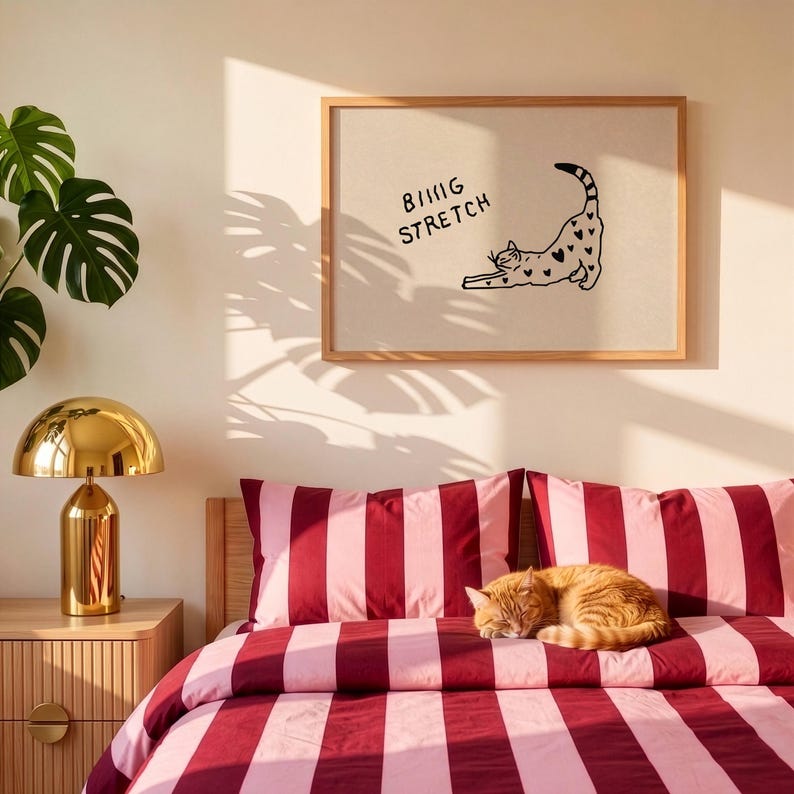 Puede incluir: Un estampado enmarcado con las palabras "Being Stretch" y una ilustraci&oacute;n de un gato con detalles de corazones. Un gato jengibre descansa sobre una cama con ropa de cama a rayas rojas y rosas. Tambi&eacute;n se ve una l&aacute;mpara dorada y una planta de monstera.