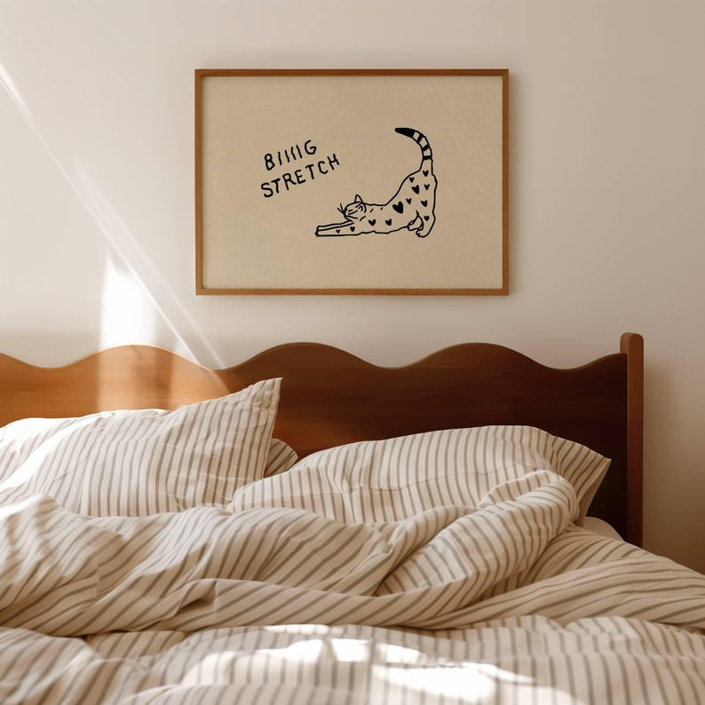 Puede incluir: Un cuadro enmarcado con un dibujo lineal negro de un gato estir&aacute;ndose con marcas en forma de coraz&oacute;n. Las palabras "BIG STRETCH" est&aacute;n encima del gato. El cuadro est&aacute; en un marco de madera, sobre una cama con ropa de cama a rayas.